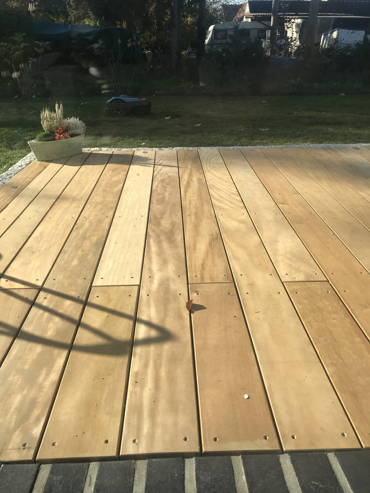 Holzdeck aus Brettern im Garten, Schatten, links Pflanzkübel mit Blumen, Gras dahinter.
