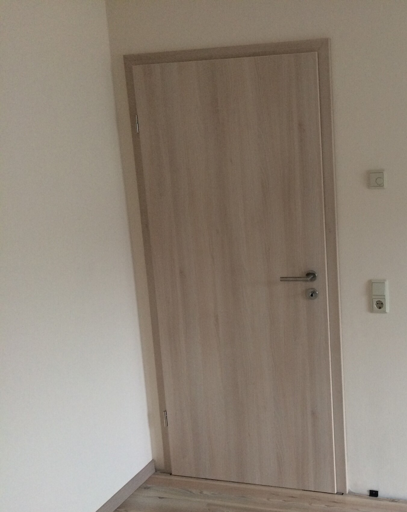Helle Holztür in weißem Raum, Türgriff rechts, Lichtschalter und Steckdose daneben.