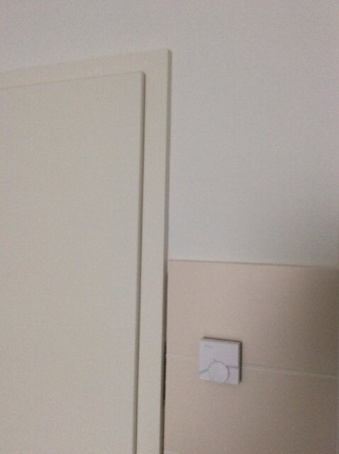 Weißer Türrahmen links, daneben kleine quadratische weiße Schalterblende auf beiger Fliesenwand.
