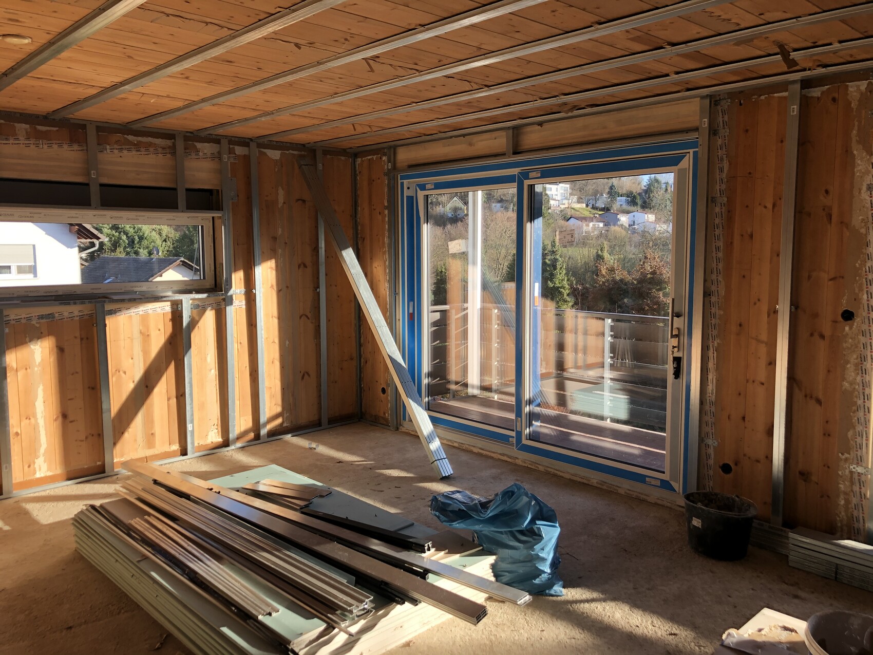 Unfertiger Innenraum mit Holzständern, Metallrahmen, Glas-Schiebetür zum Balkon, Baumaterialien.
