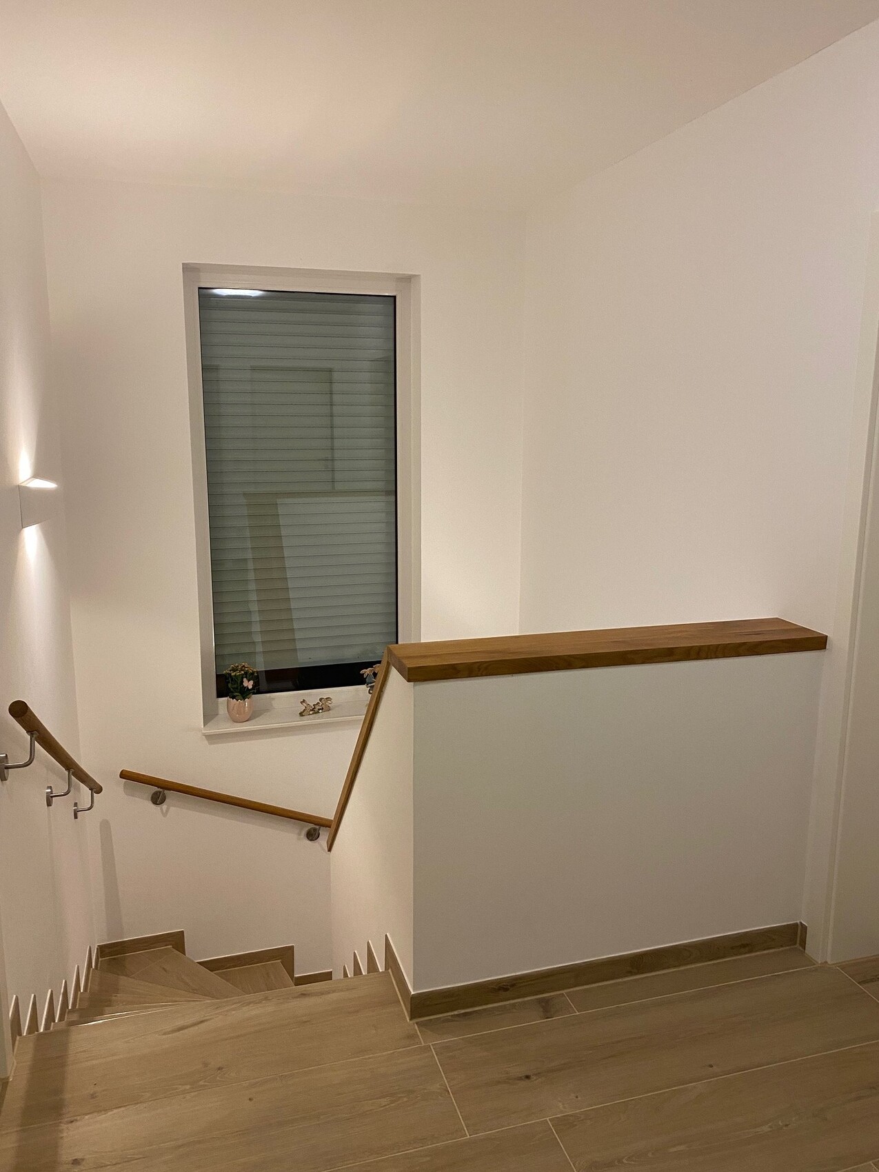 Heller Treppenaufgang mit Holzgeländer, Fenster mit Jalousien am Absatz, weiße Wände, Holzboden.