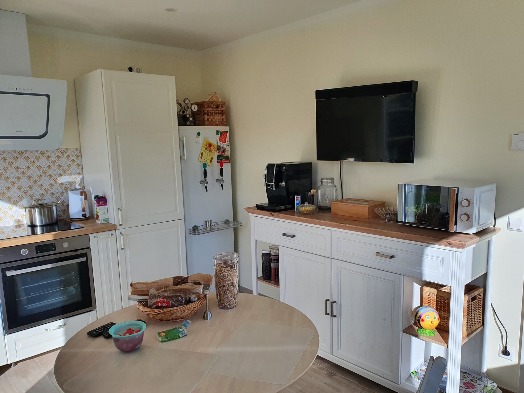 Küche mit weißen Schränken, Kühlschrank, Backofen, Mikrowelle, Kaffeemaschine, Wand-TV und Esstisch.