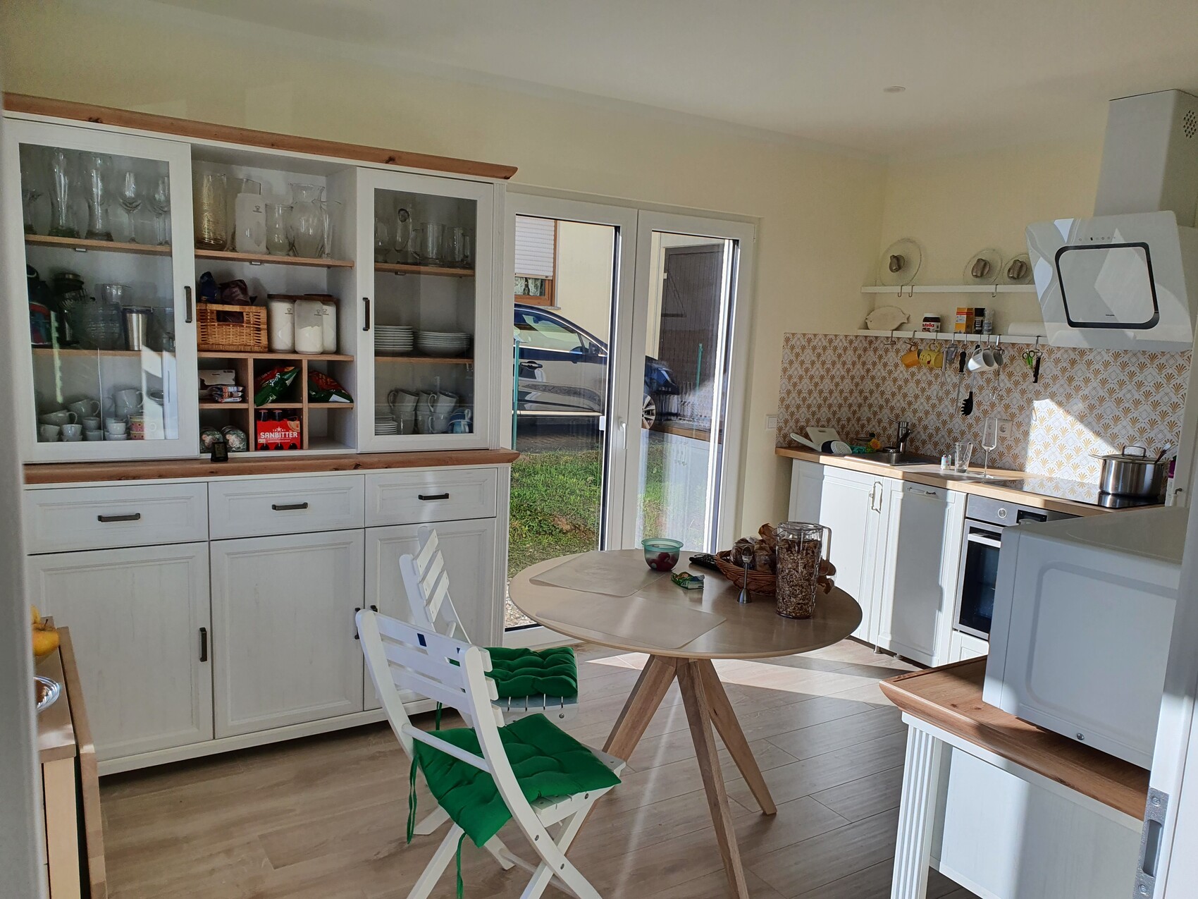 Helle Küche mit weißen Schränken, Glasvitrinen, rundem Holztisch und grünen Sitzkissen.