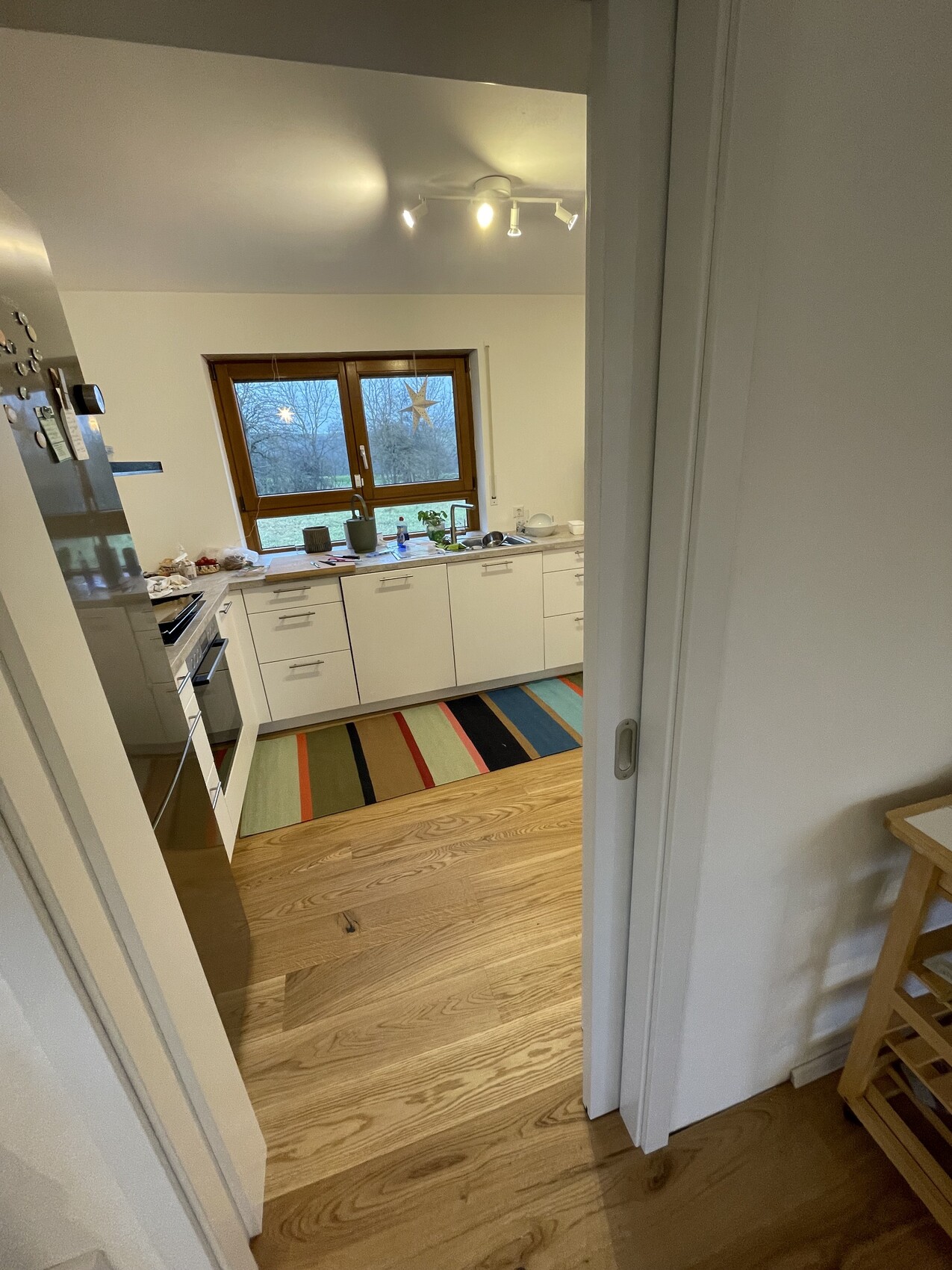 Küche mit weißen Schränken, Holzboden, bunter Streifen-Teppich, Fenster mit Sternen.