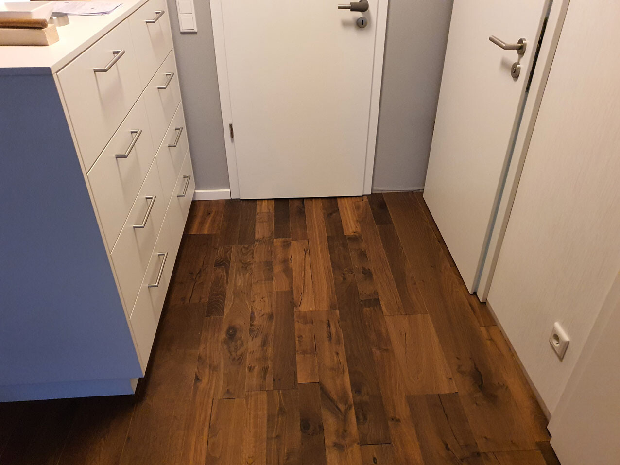 Küchenflur mit weißen Schubladen links, blaues Seitenpanel, dunkler Holzboden, Türen rechts.