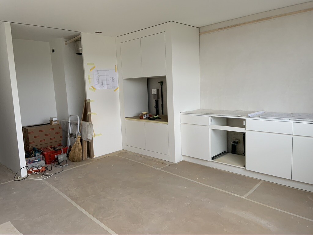 Küche im Bau: weiße Schränke, Arbeitsbereich; links Kisten, Leiter, Reinigungswerkzeug.