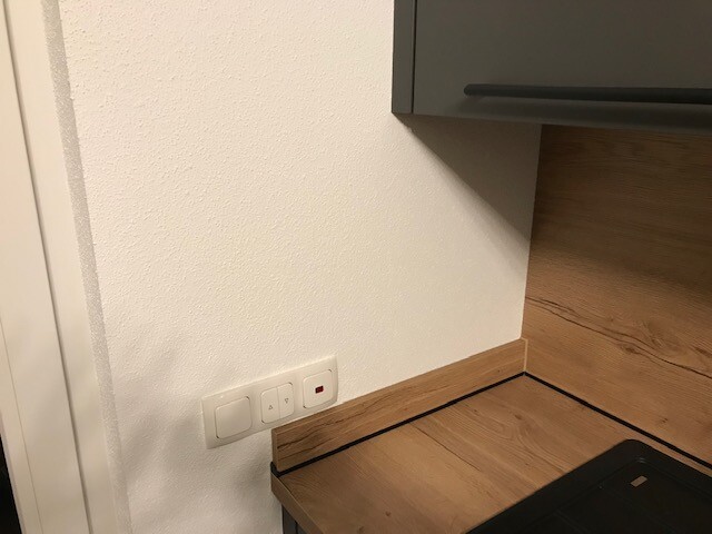 Küchenkante mit Holzarbeitsplatte, weißer Wand, grauem Oberschrank und Steckdosenplatte.