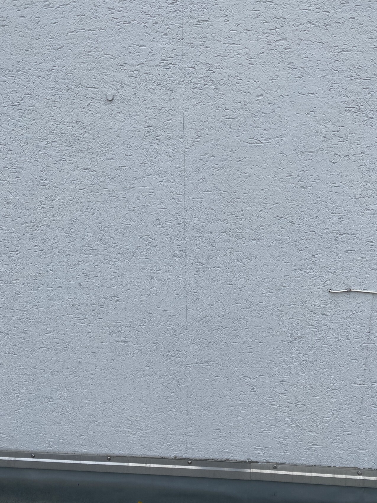 Graue, raue Wand mit senkrechter Fuge, kleinem Knopf und seitlichem Kabel an der Seite.