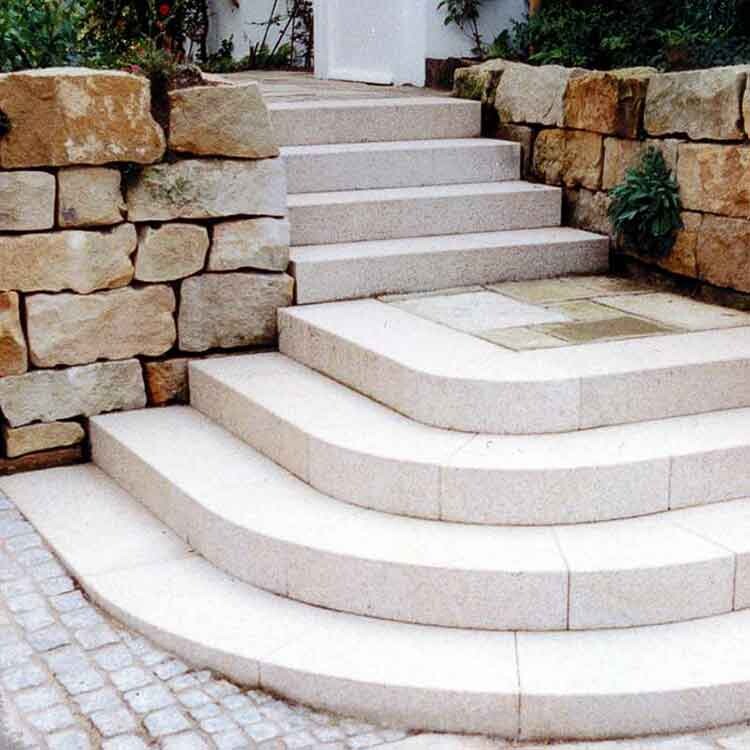 Helle Naturstein-Treppe mit halbrunden Stufen, flankiert von Sandsteinmauern, führt zu einer Tür.