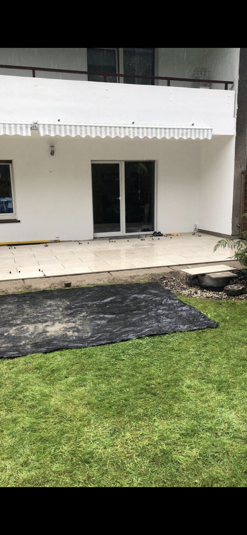 Terrasse vor einem weißen Haus mit Glas-Schiebetür; schwarze Abdeckplane auf dem Gras, Bauarbeiten