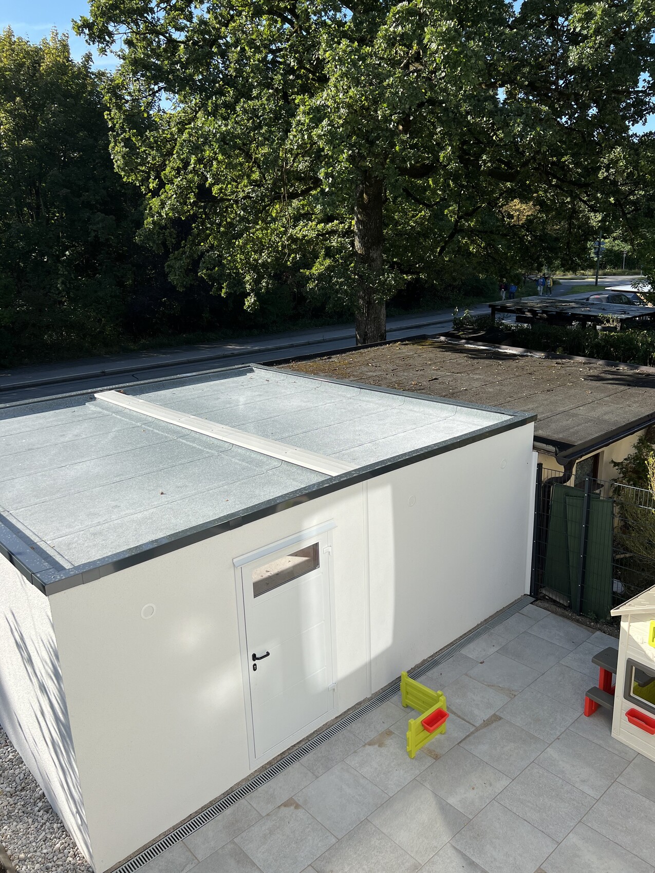 Kleines weißes Gebäude mit Flachdach; Terrasse mit buntem Kinderspielzeug, Bäume dahinter.