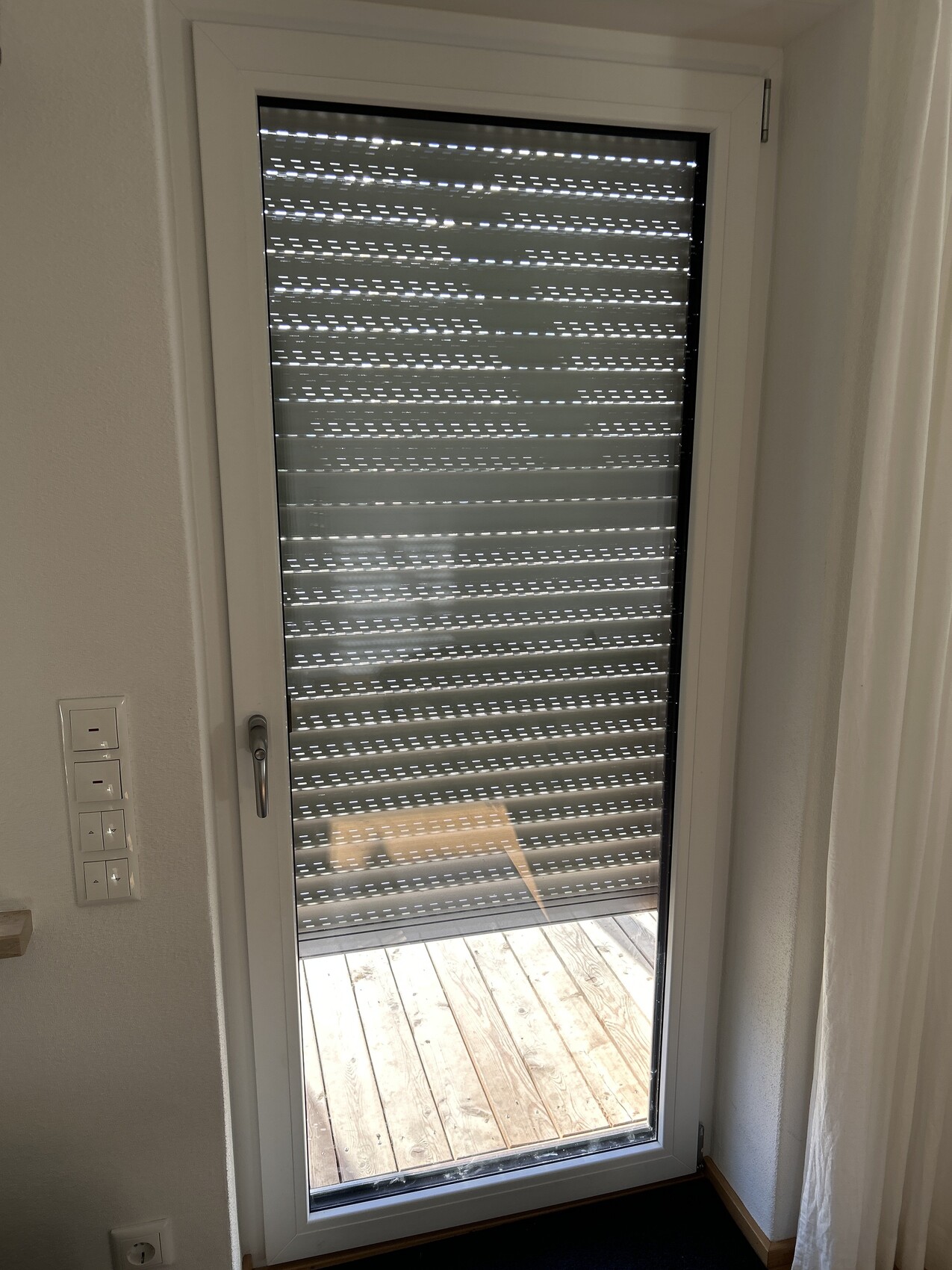 Glas-Schiebetür mit Rollladen, teils geöffnet, Blick auf Holzdeck draußen.