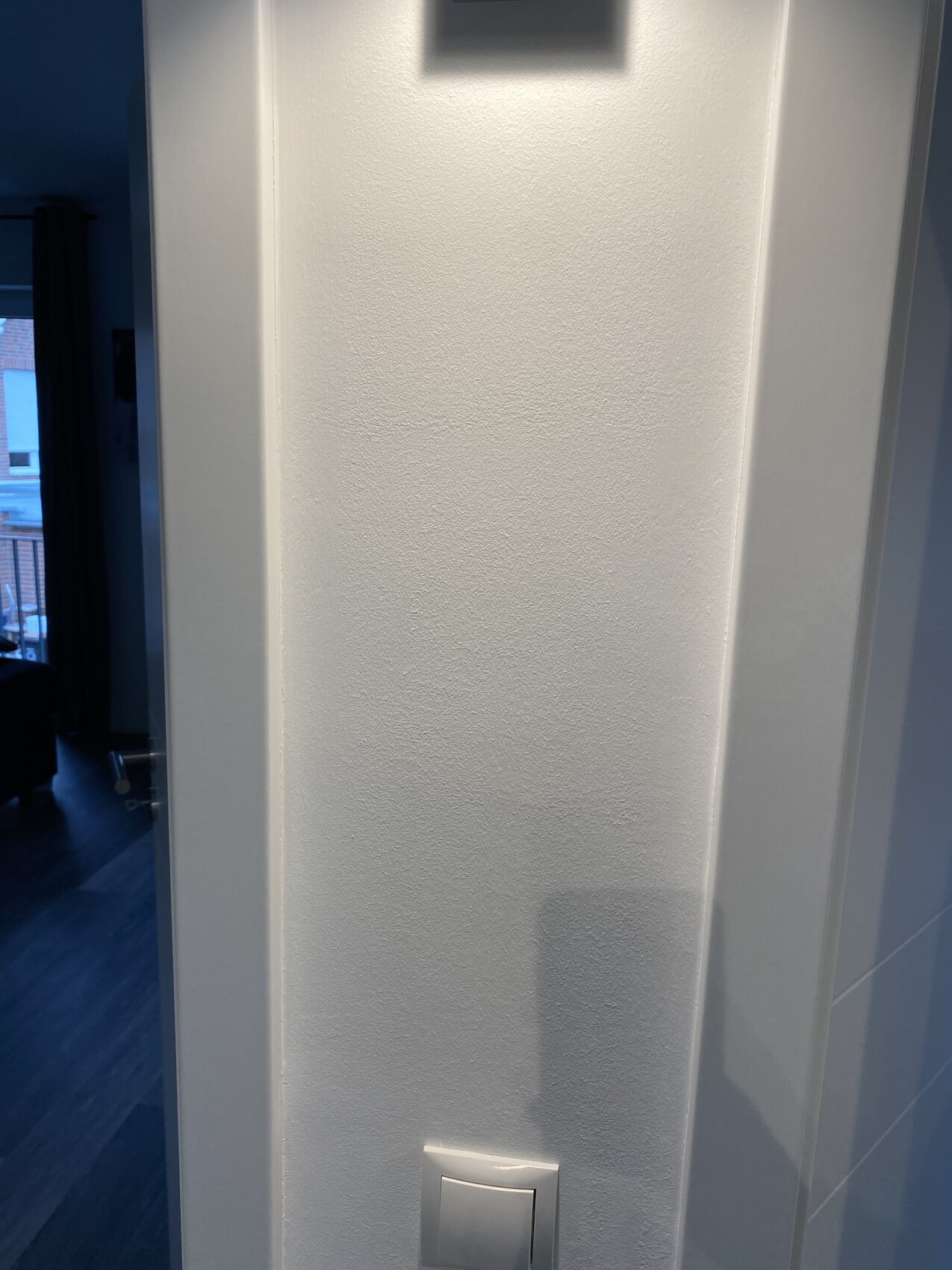 Weiße texturierte Wand mit kleinem Gerät oben und unten Lichtschalter; linker Durchgang sichtbar.