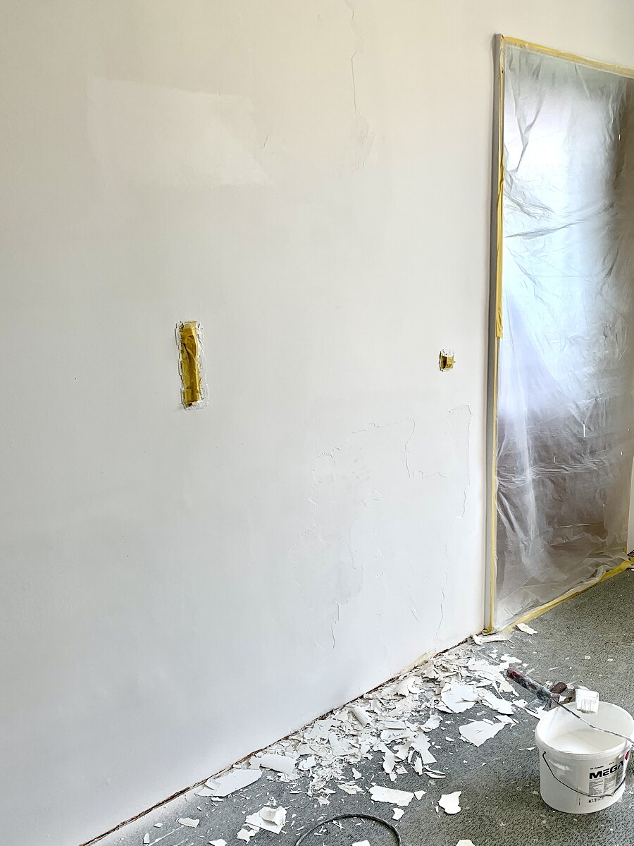 Renovierungsraum: Weiße Wand mit Putzrissen, Türbereich hinter Plastikfolie, Boden mit Trümmern.