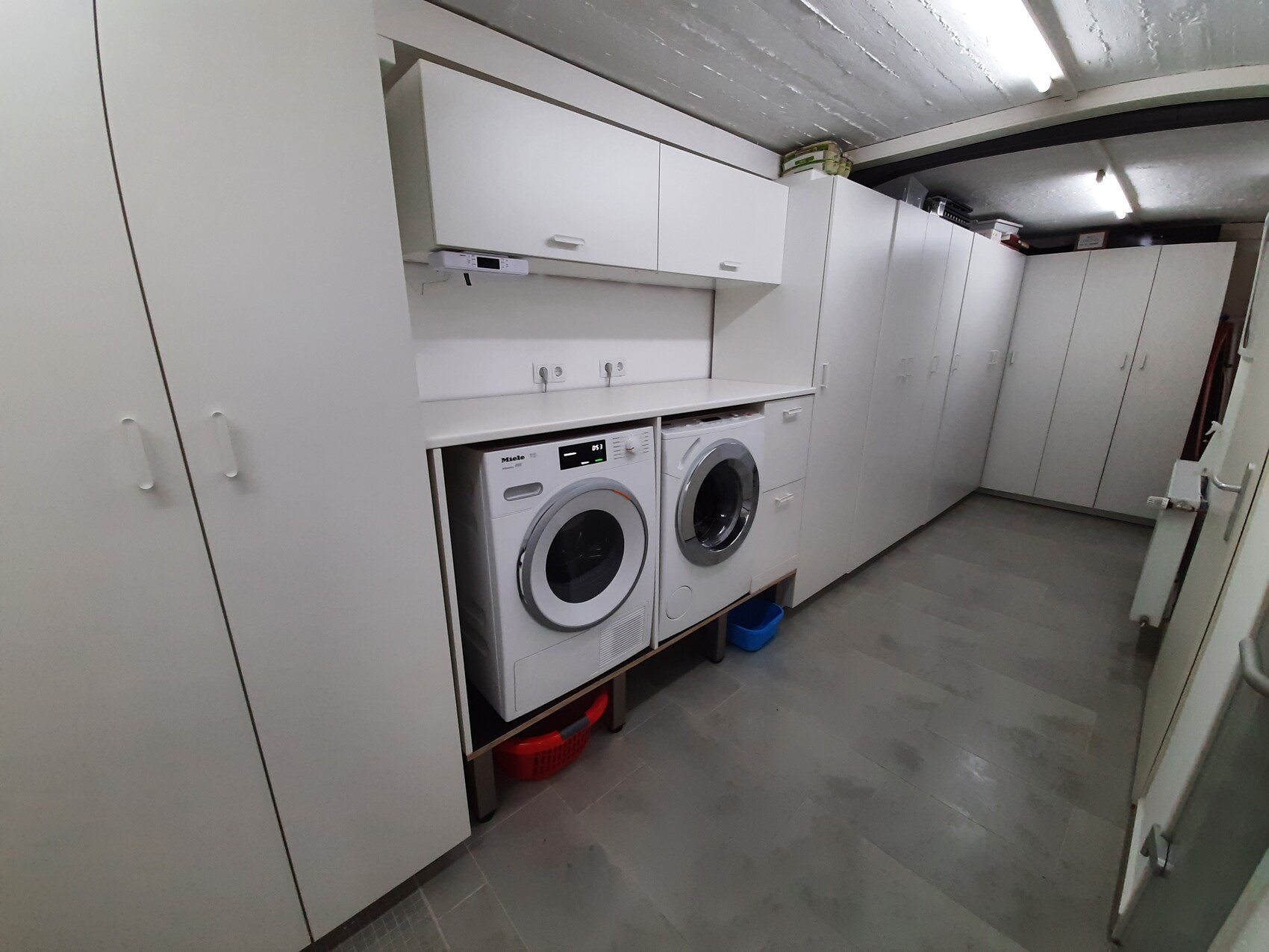 Waschküche mit zwei Frontlader-Waschmaschinen unter einer weißen Arbeitsplatte und vielen Schränken.