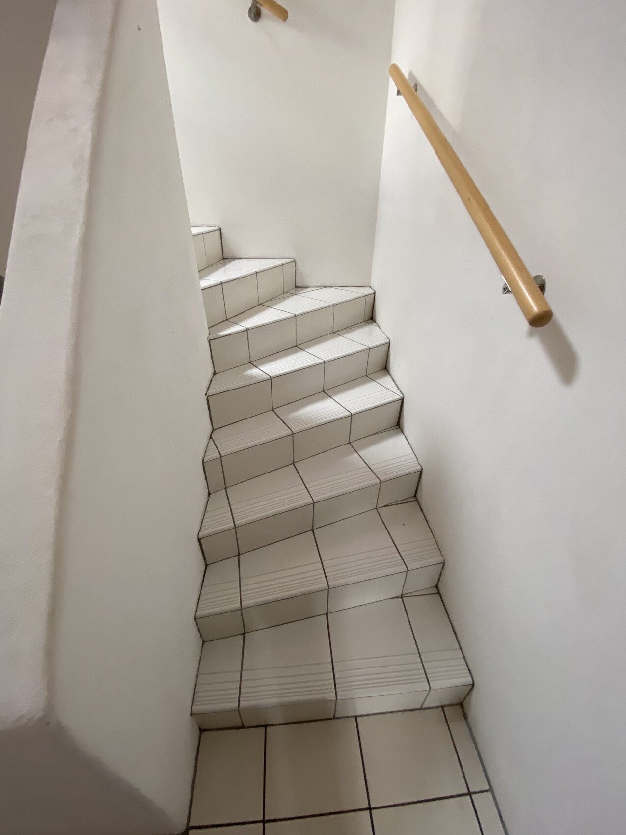 Weiße Fliesenstufen einer engen Treppe, Holzhandlauf rechts, Innenraum-Treppenaufgang.