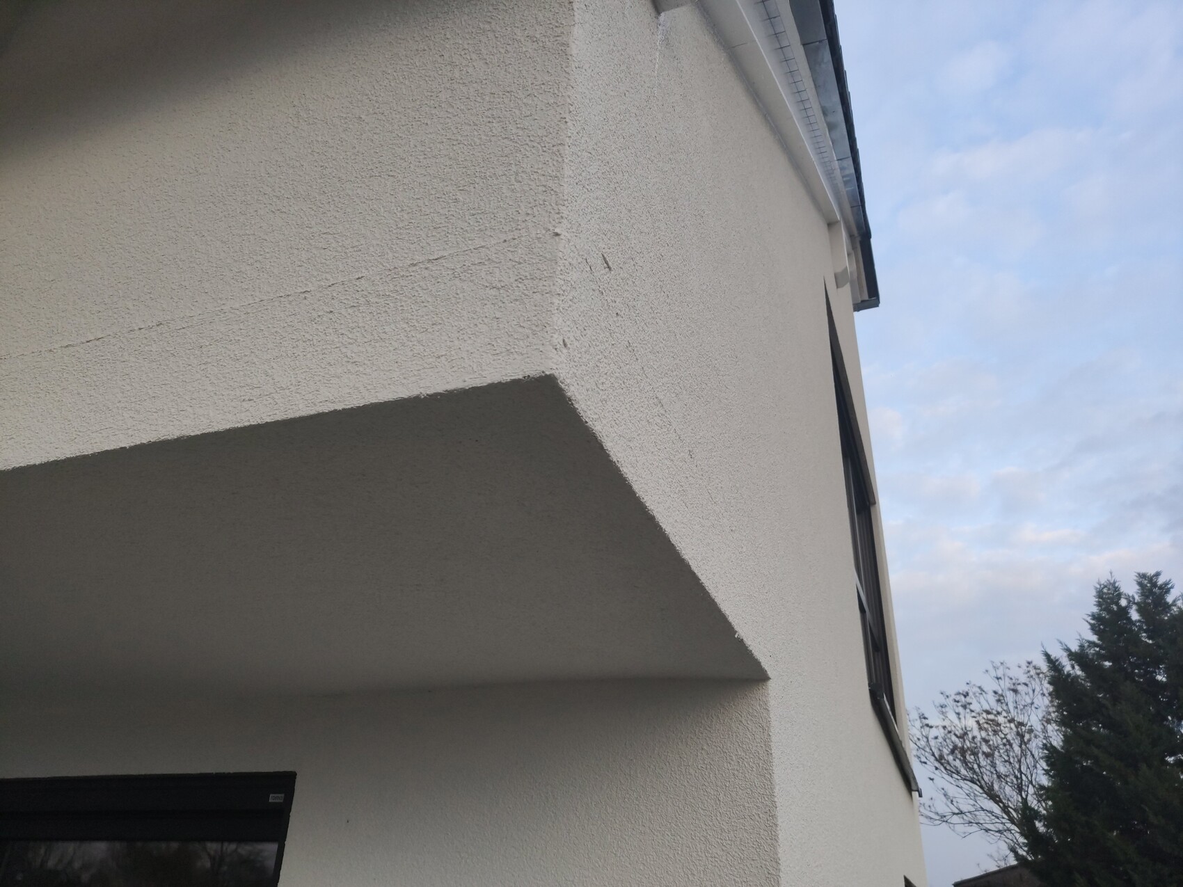 Weiße Gebäudefassade mit schrägem Überhang, Fenster unten links, blauer Himmel rechts.