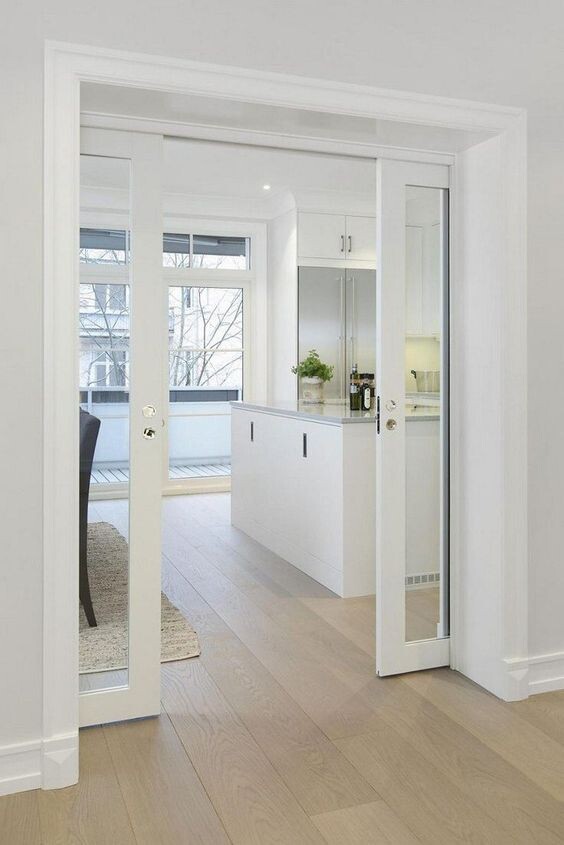 Offene weiße Glastüren führen in eine helle Küche mit Insel, Fensterfronten und Holzboden