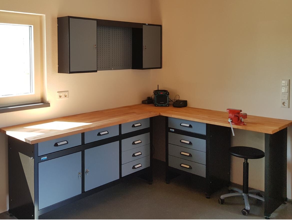 Werkstatt-Ecke mit Holz-Arbeitsplatte, grauen Schubladen, Wandschrank und Fenster links.