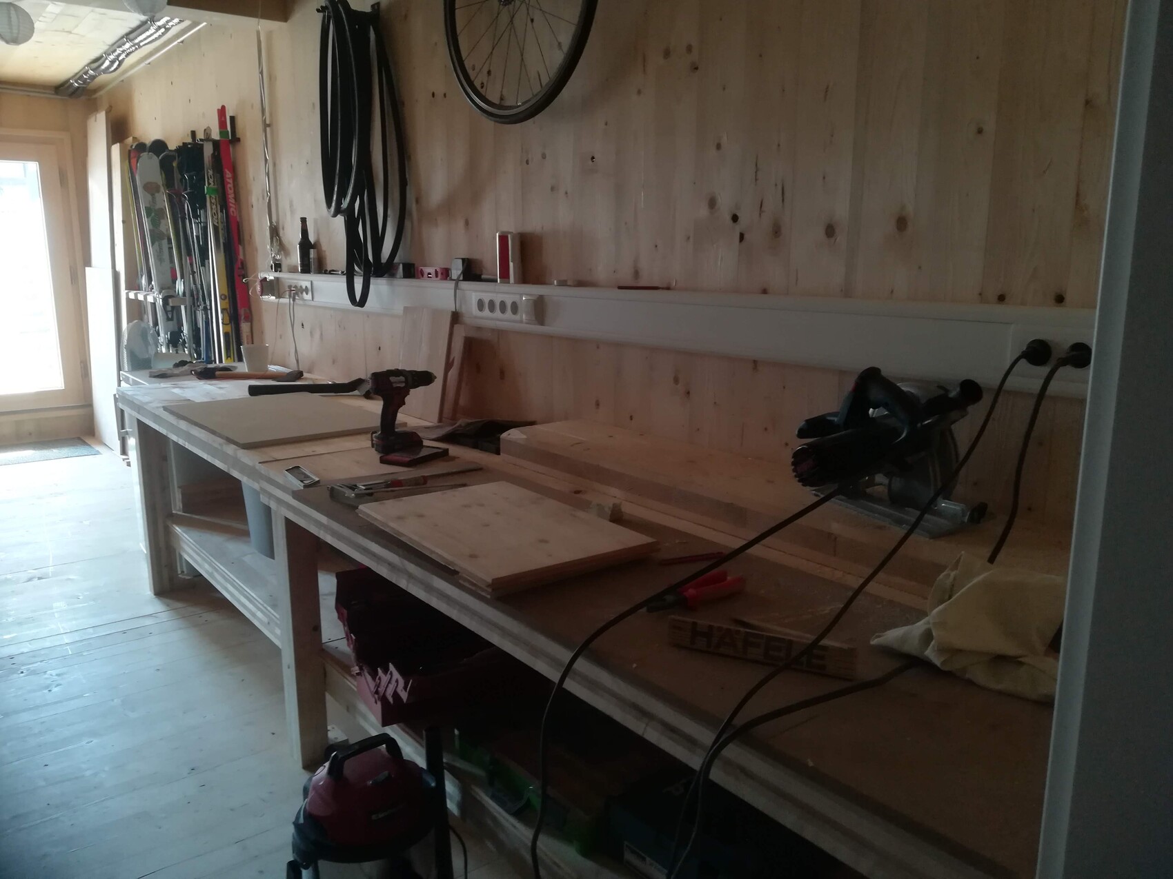 Holzwerkstatt mit Werkbank, Bohrmaschine, Säge und Holzplatten auf der Arbeitsfläche.