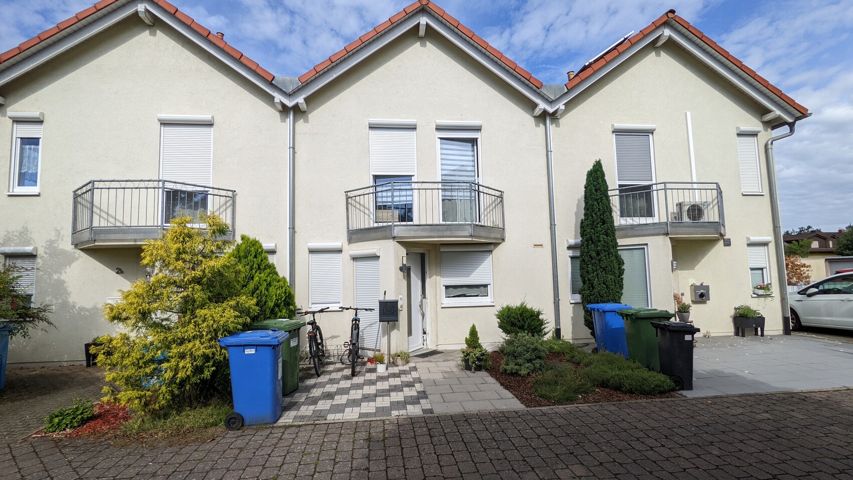 Beiges Mehrfamilienhaus mit Balkonen, Gartenpflanzen, Mülltonnen und Fahrrad am Eingang.