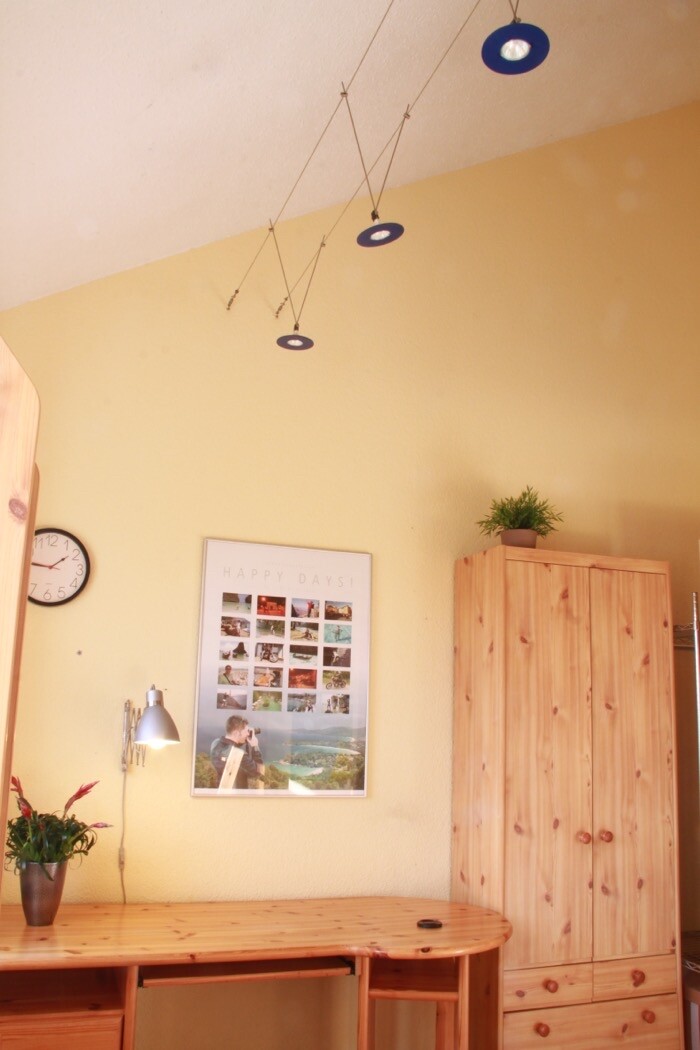 Heller Raum mit gelber Wand, Holzschreibtisch, Kleiderschrank, Pflanze, Uhr und Poster 'Happy Days'.