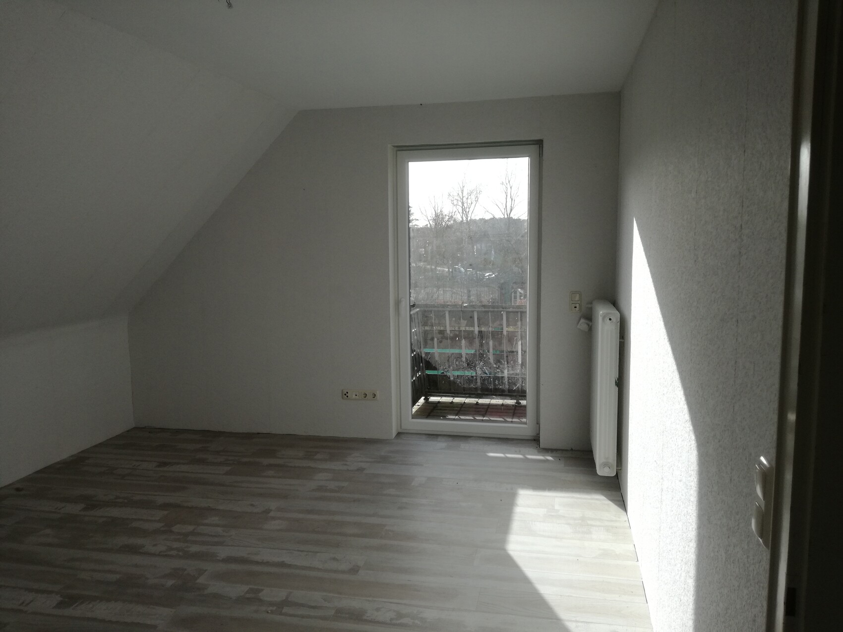 Leerer Dachbodenraum mit schräger Decke, Holzfußboden, Tür zum Balkon, Heizkörper an der Wand.