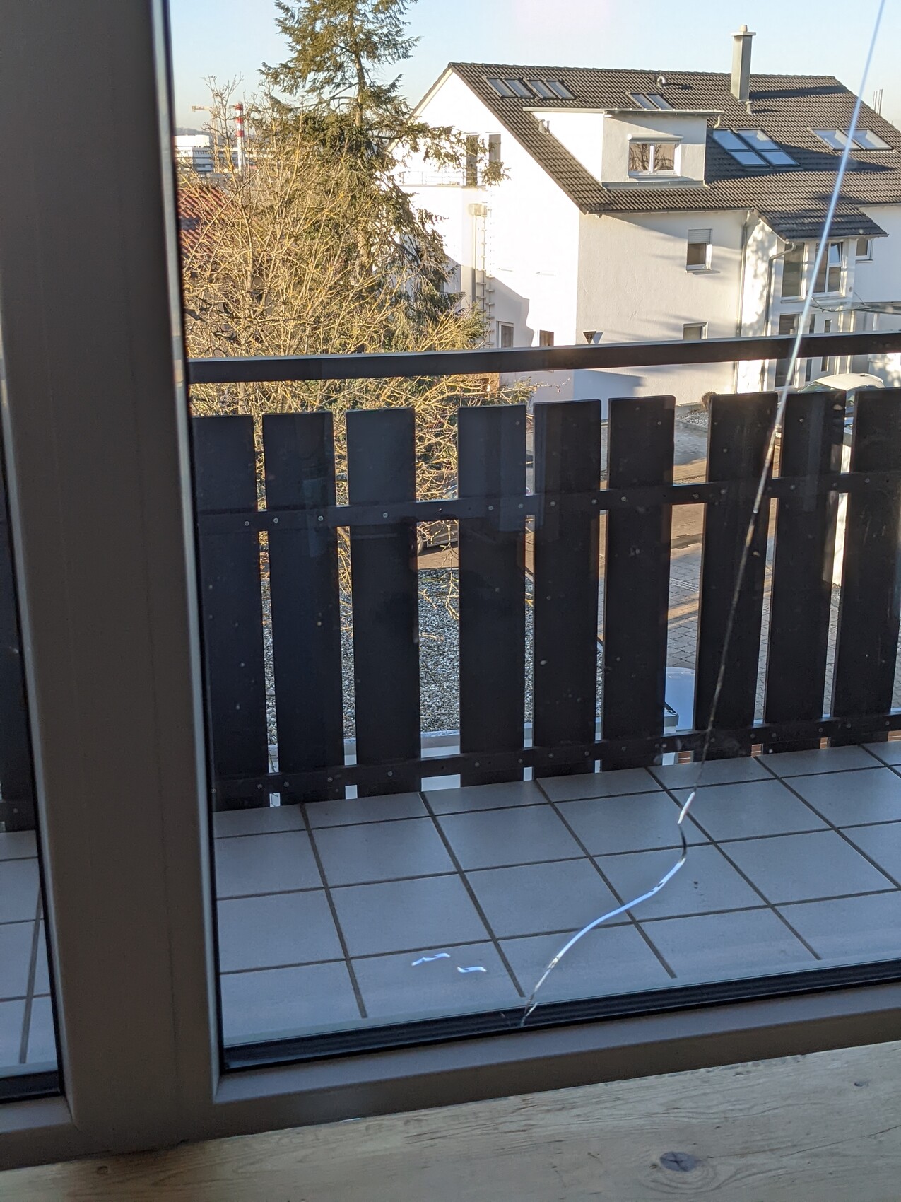 Blick durch Glasfenster auf Balkon mit dunklem Holzgeländer; Häuser, Baum, Fliesenboden.