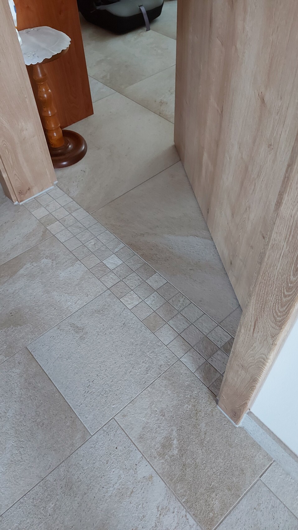 Beige Fliesenboden mit schmaler Quadratbordüre; Holz-Türenrahmen rechts, Tischbein im Hintergrund.