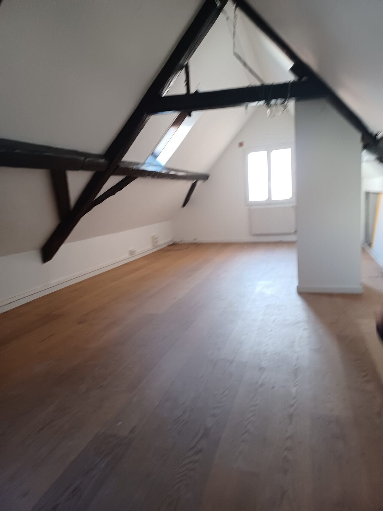 Dachgeschoss mit dunklen Holzbalken, heller Holzboden und Fenster am Ende des Raums.