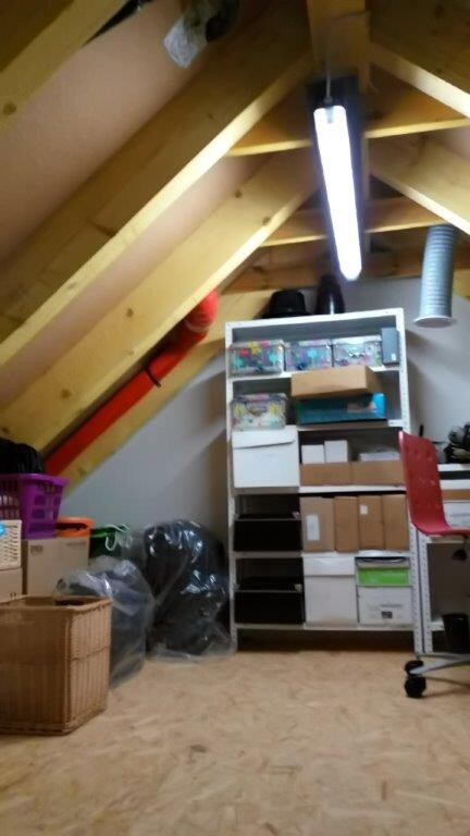 Dachboden mit Holzbalken, Regalen, Kartons und Aufbewahrungsboxen unter einer Lampe.