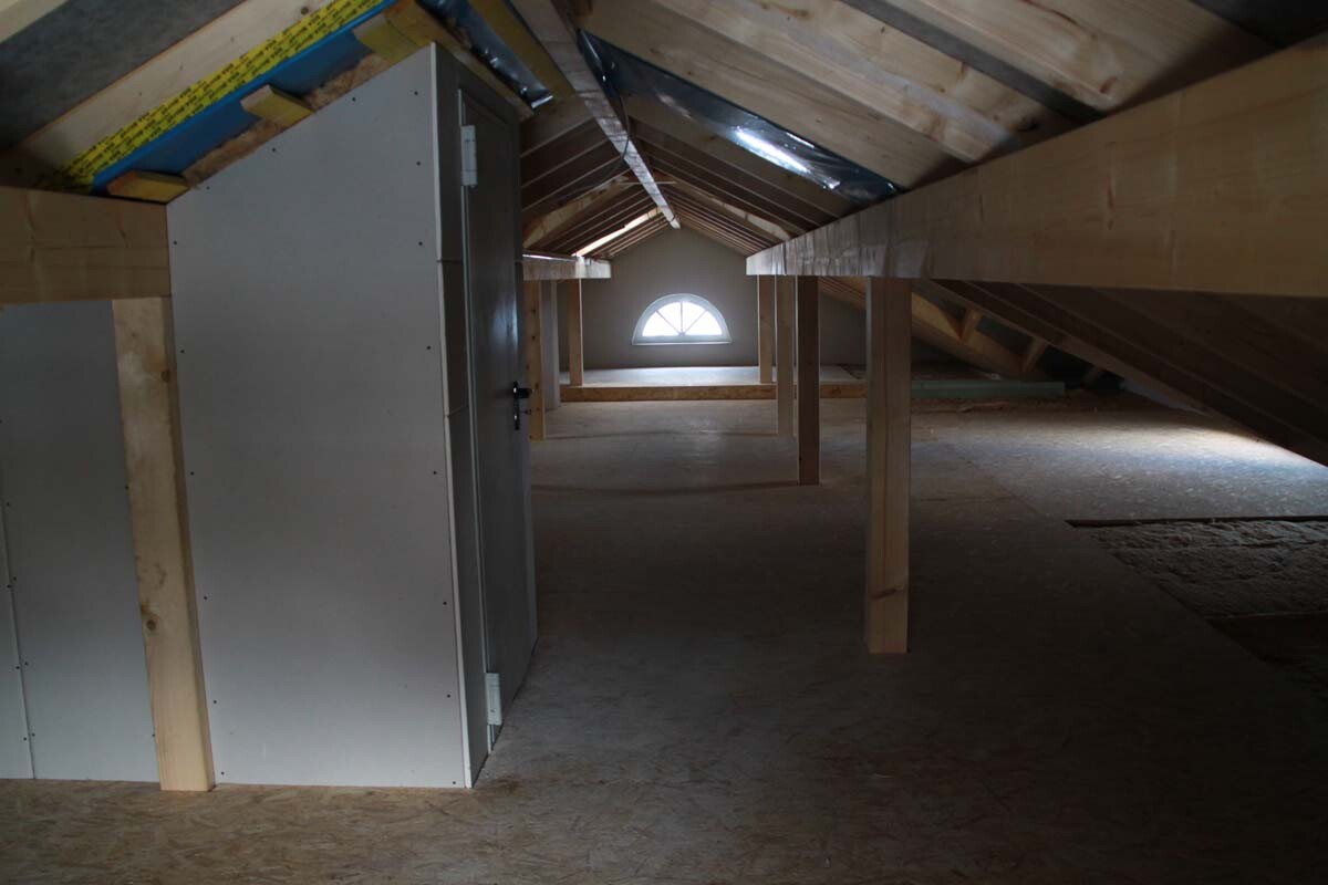 Dachboden mit Holzbalken, links steht ein großer Metallschrank, hinten ein Fenster.