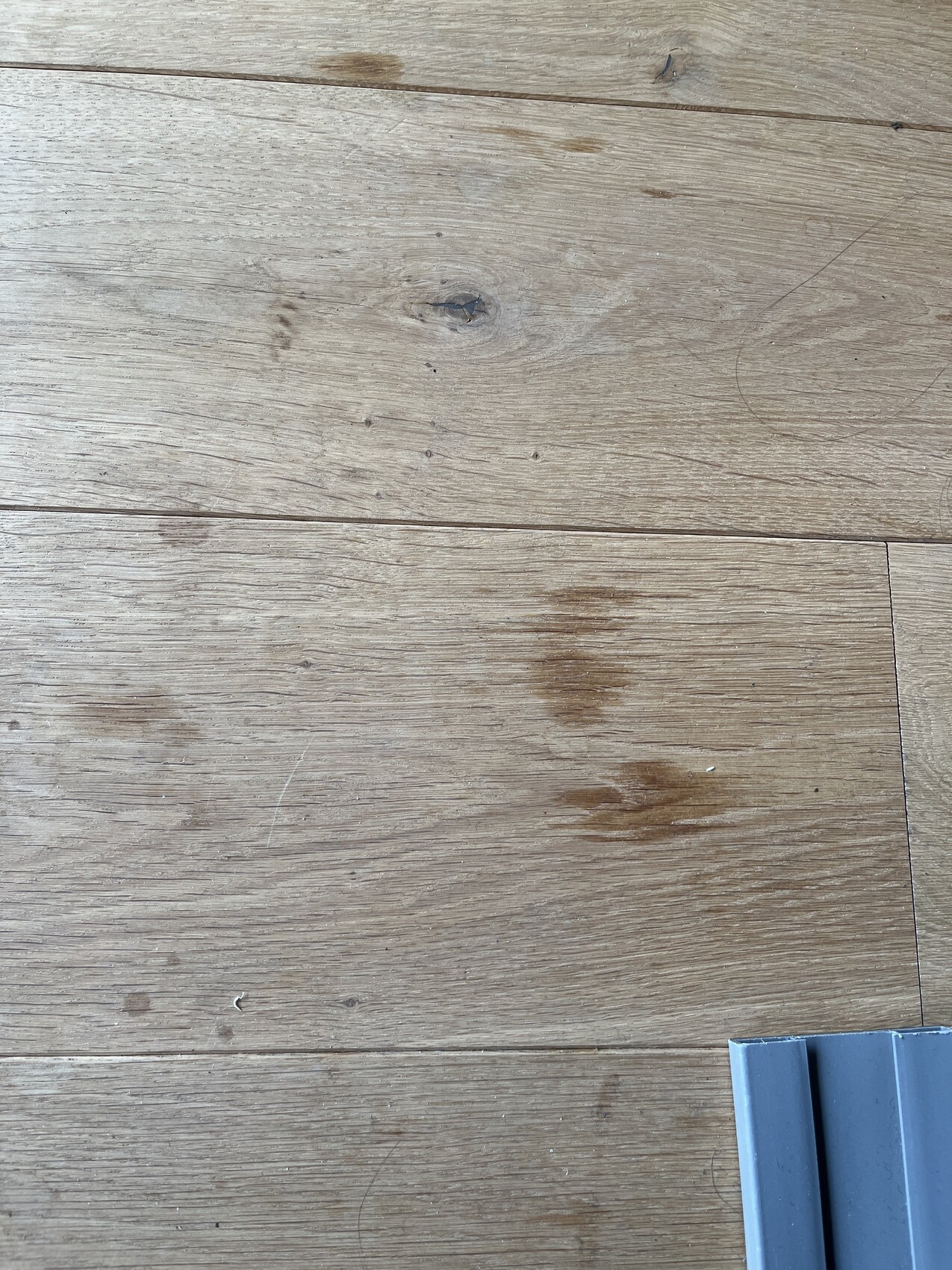 Holzboden mit Flecken; sichtbar ein graues Metallteil unten rechts.