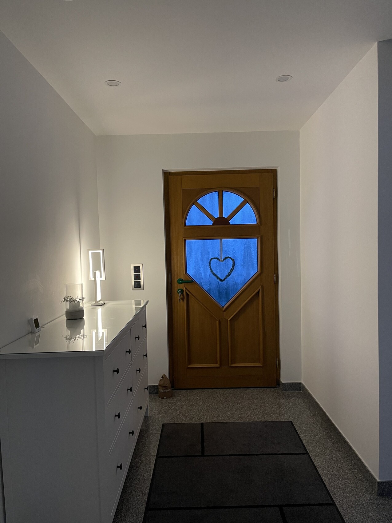 Heller Flur mit Holztür, Glaseinsatz mit Herz, weiße Kommode, dunkler Teppich.