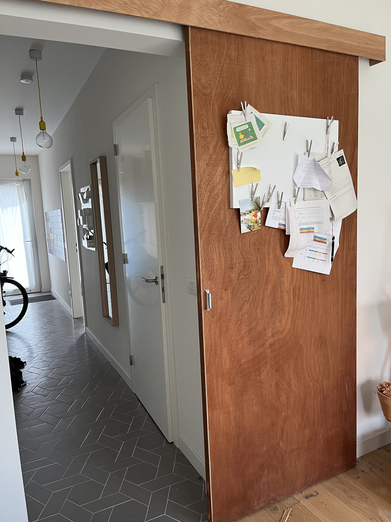 Heller Flur mit grauem Fliesenboden, Holzwand mit Pinnwand voller Zettel; links Fahrrad.