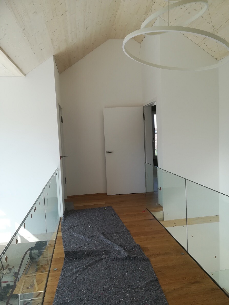 Heller Flur mit Holzboden, Glasgeländer, weißen Wänden und runden Deckenleuchten.