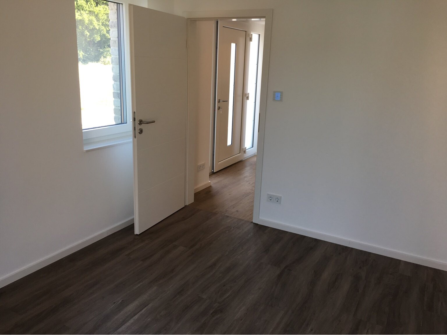 Leerer Raum mit dunklem Holzboden, offener Tür zum Flur und Fenster an der linken Wand.