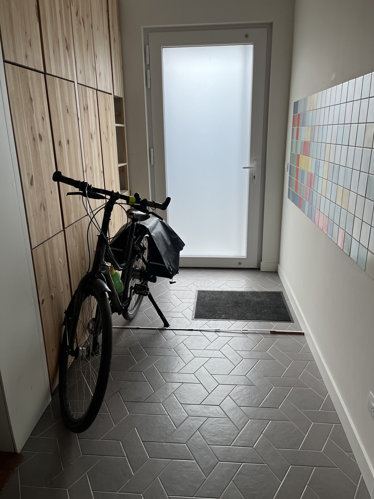 Fahrrad steht im Flur vor Holzschränken, Tür mit milchigem Glas, bunte Wandfliesen rechts.