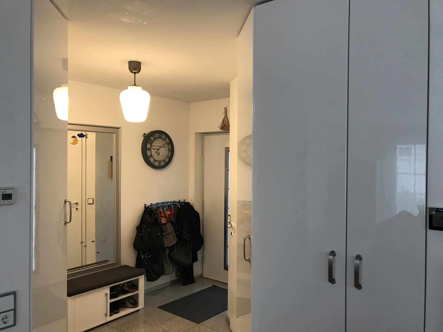 Heller Eingangsflur mit Spiegel, Uhr, Garderobe mit Taschen, Schuhbank und Tür.