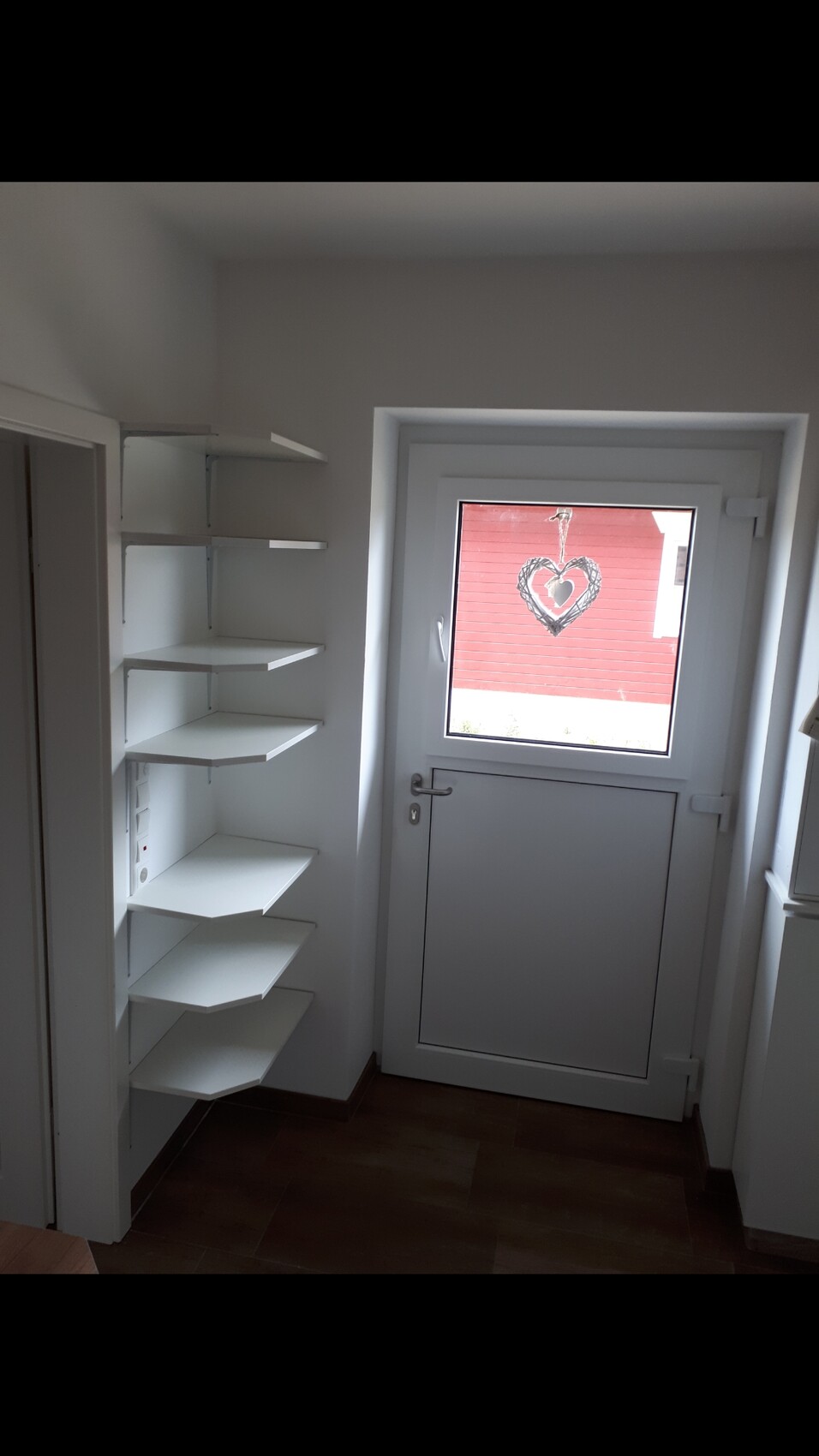 Flur-Ecke mit weißen Eckregalen links, Tür mit Fenster, draußen hängt eine Herz-Deko.