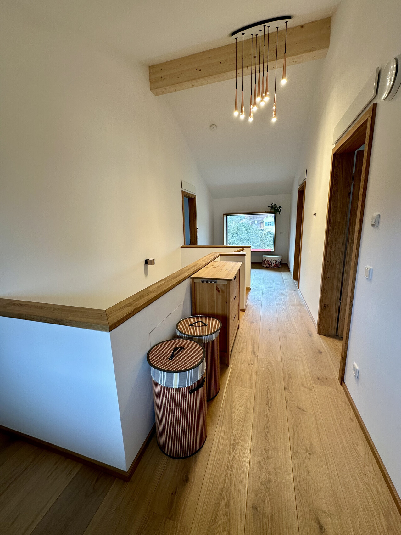 Heller Flur mit Holzboden, Holzbalken, Pendelleuchten, zwei runde Wäschekörbe, Fenster am Ende