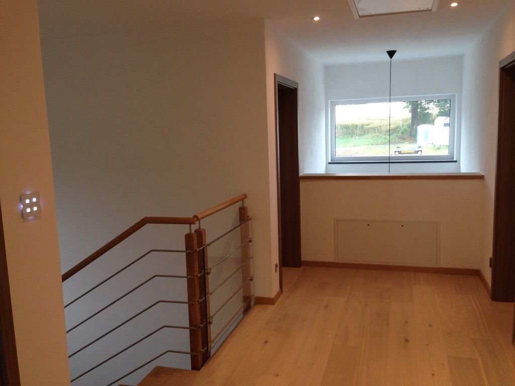 Heller Flur mit Treppe links, Geländer aus Holz und Metallseilen, Fenster mit Blick nach draußen.
