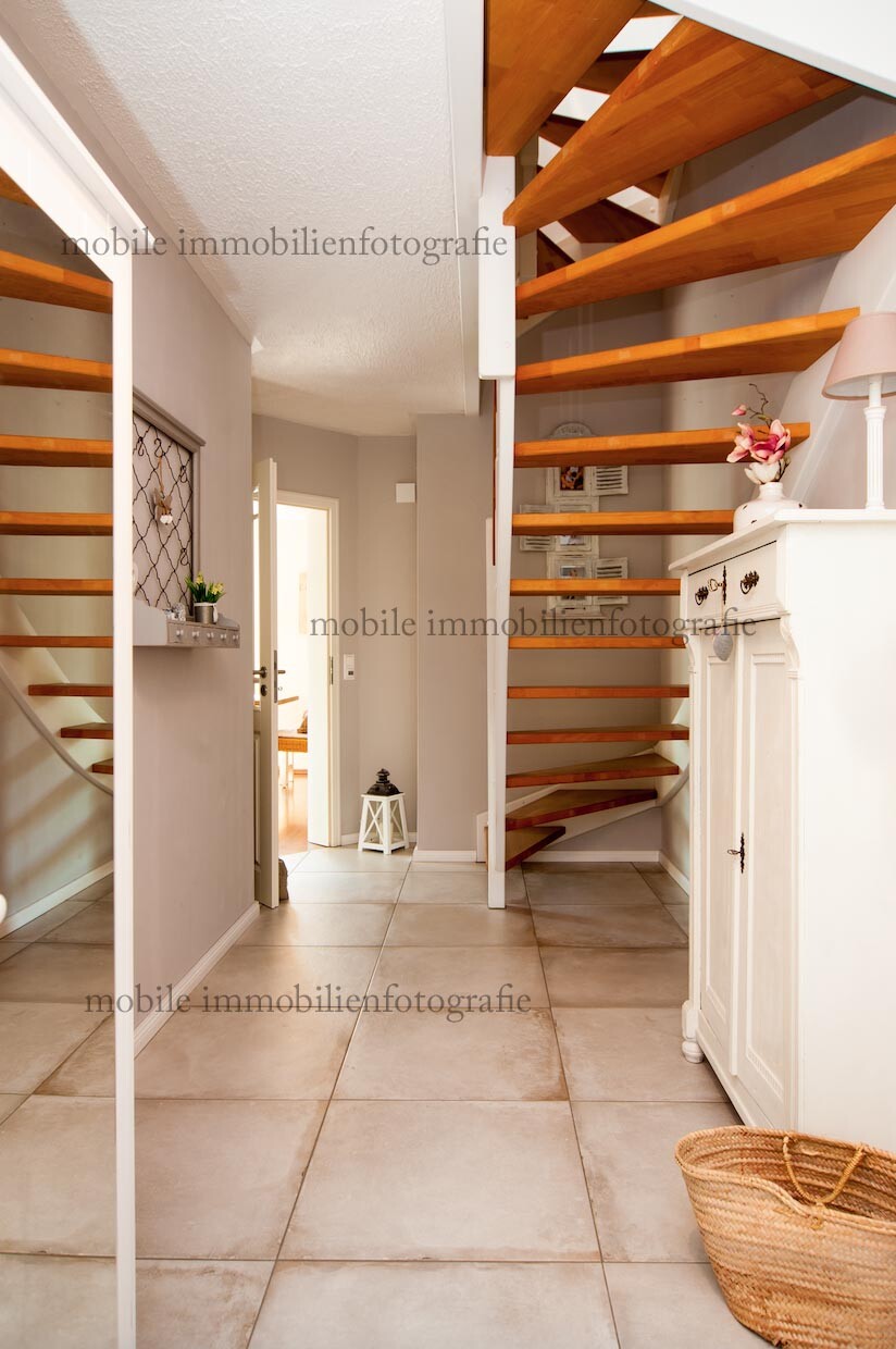Hell beleuchteter Flur mit offener Holztreppe rechts, weißen Möbeln und Tür im Hintergrund.