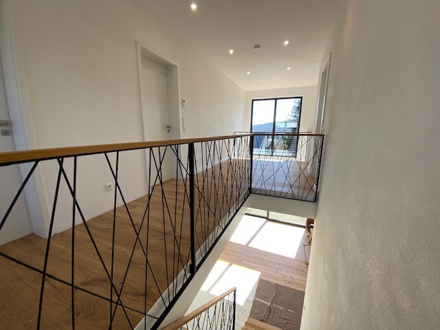 Heller Innenflur mit Holzboden, Metallgeländer mit Muster, Treppe nach unten und Fenster am Ende.