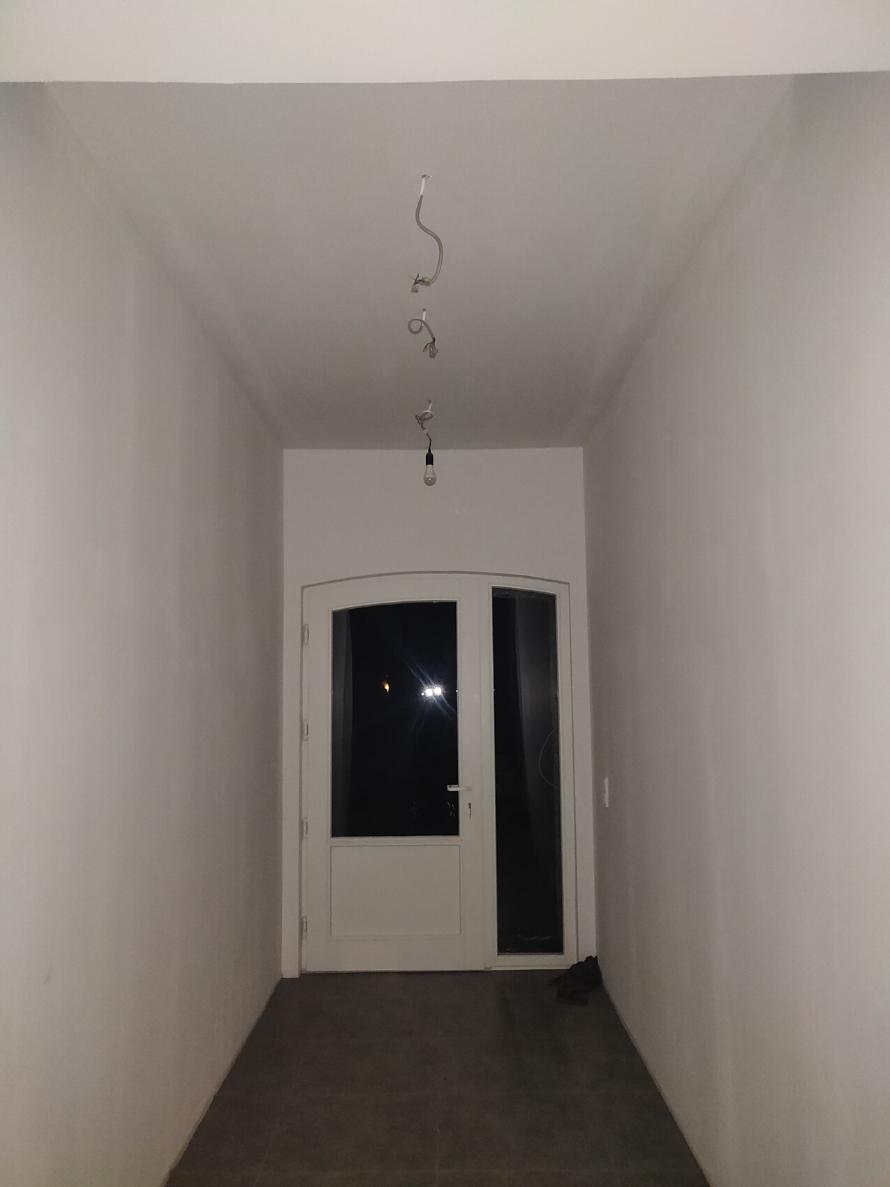 Langer Flur mit weißen Wänden, dunklem Boden; Tür am Ende mit Glasfenstern.