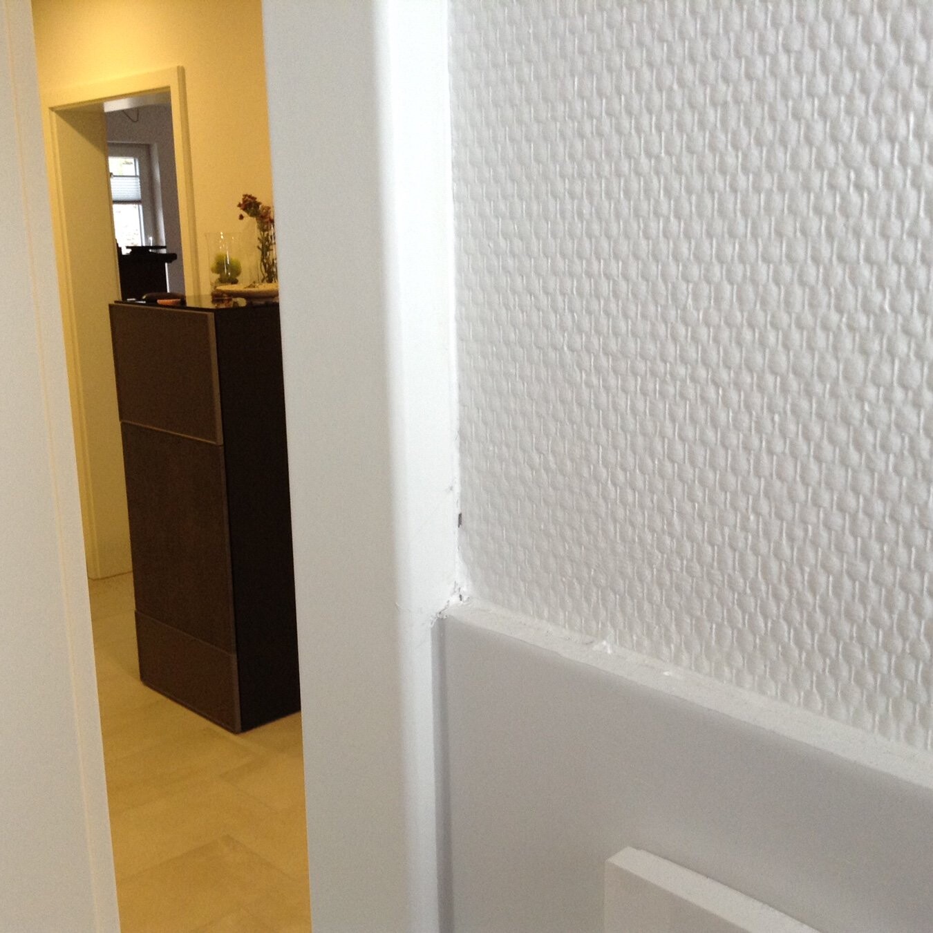 Blick in einen Flur: weiße strukturierte Wand rechts, dunkler Schrank links, Türrahmen im Vordergrund.