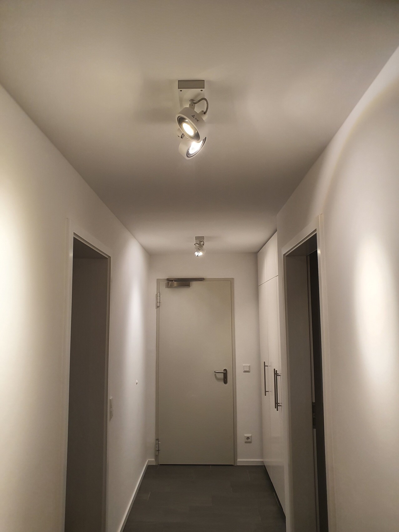 Heller Flur mit weißen Wänden, grauem Boden, Türen links und rechts, Tür am Ende, Deckenstrahler