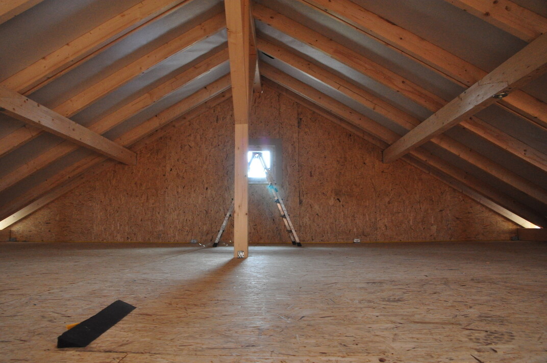 Dachboden mit Holzbalken, OSB-Wänden, zentrale Säule, Leiter zum Fenster, Licht von außen.