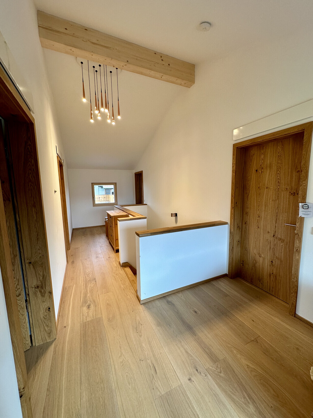 Heller Flur mit Holzboden, einem Holzbalken an der Decke, hängenden Leuchten und Holztüren