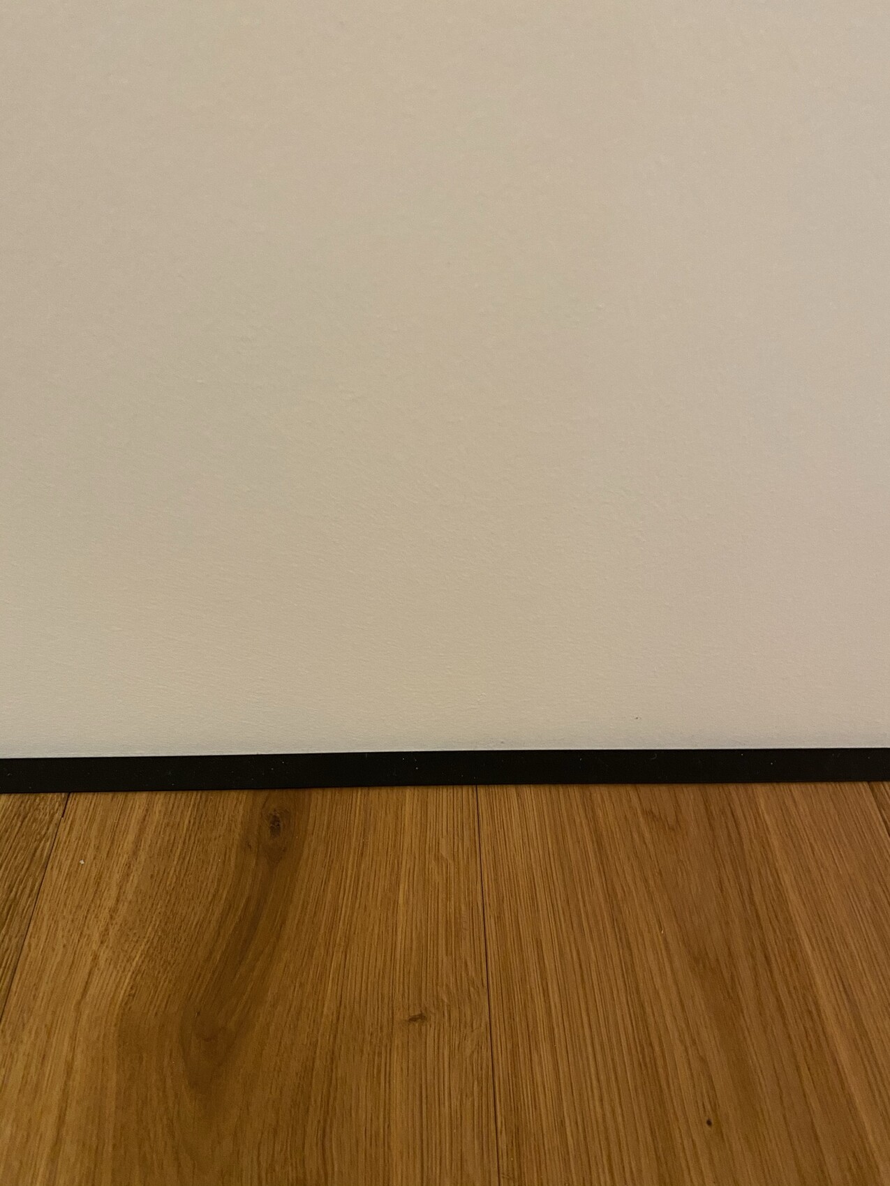 Weiße Wand mit schwarzer Sockelleiste über braunem Holzboden.